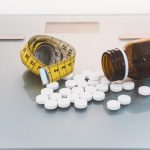metformine-weight-loss-and-diet-pills-spilling-out-of-glass-bottle-next-to-measuring-tape-on-top-of-weighing-scales
